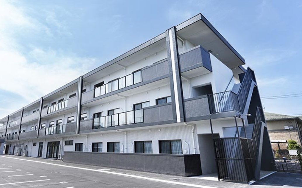 モダンデザインの3階建て介護施設。白と濃灰色の外壁、複数のバルコニーが特徴。