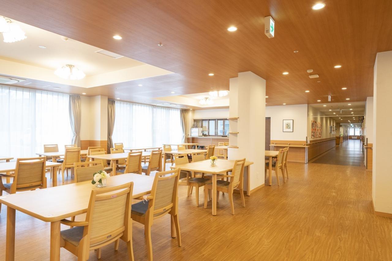 木質調の天井と明るい採光が特徴の食堂。複数のテーブルと椅子が配置され、奥には厨房が見える。