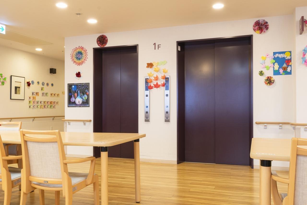 木製の椅子とテーブルがある明るい共用ロビー。壁面に入居者の作品や装飾が飾られている。