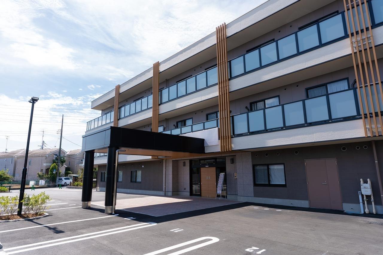 介護施設の建物外観。モダンな3階建て構造で、玄関ポーチと広い駐車場を備えた新築施設。