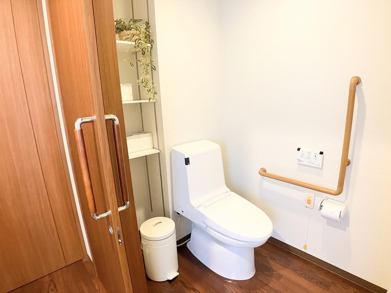 介護施設の浴室・トイレ。温水便座付きの洋式トイレと手すり、観葉植物が配置されています。